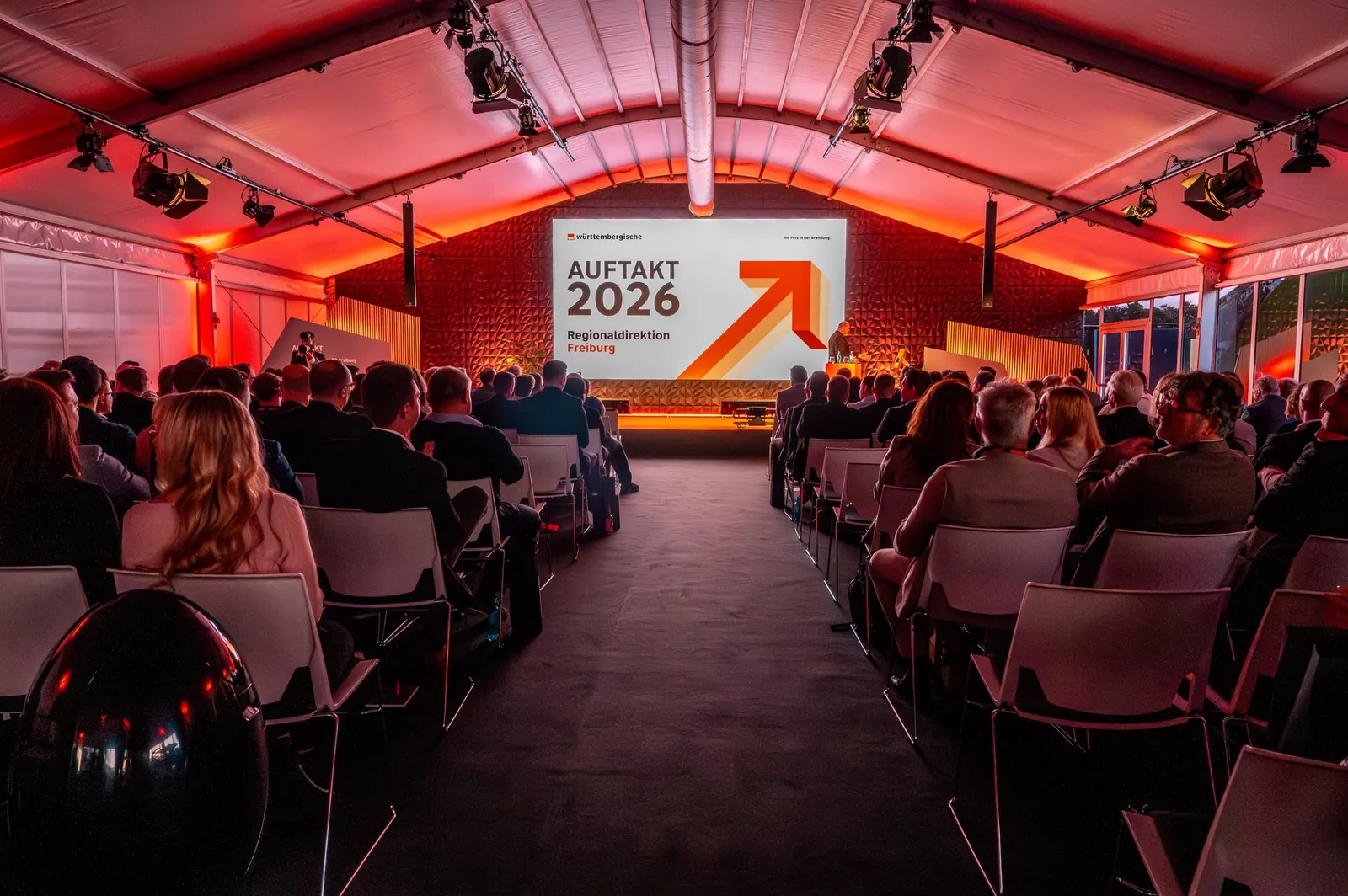 Tagung im Glaspalast: Stuhlreihen vor einer Bühne mit der Aufschrift Auftakt 2026 in rotem Licht