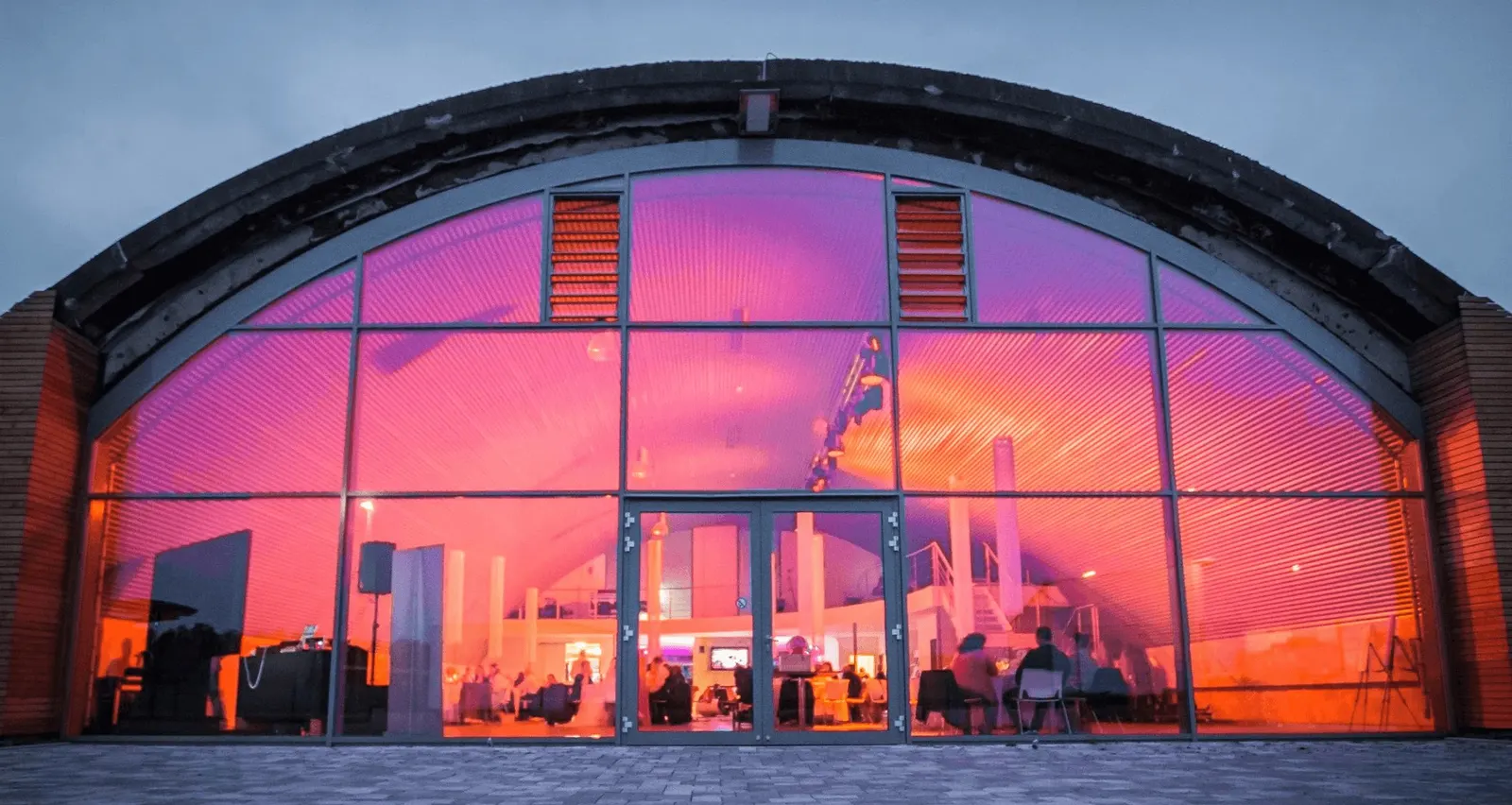 Glasfront des Hangars in der Dämmerung, innen in rotes und violettes Licht getaucht